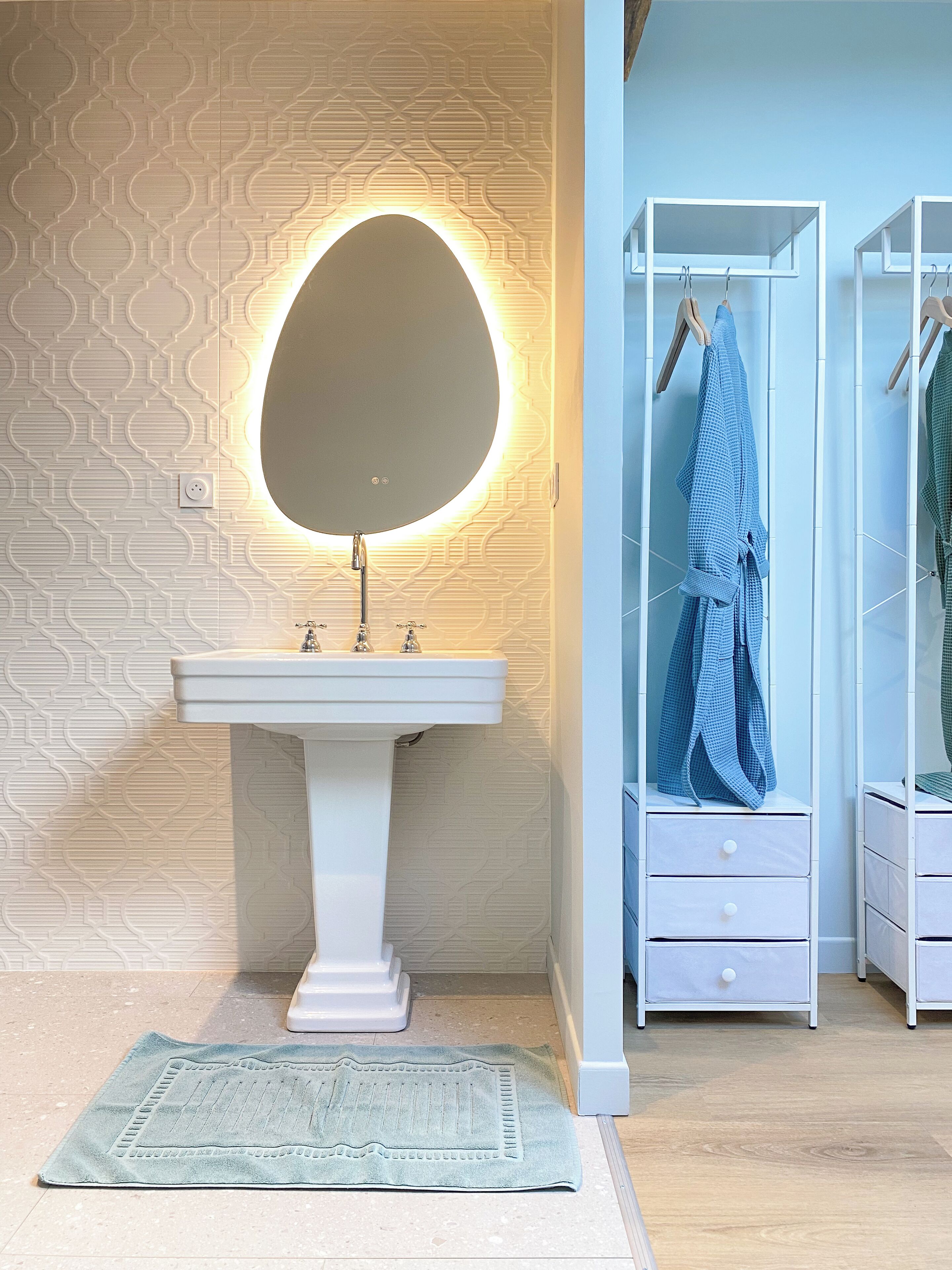 Un baño con un lavamanos blanco de pedestal, un espejo redondo con iluminación trasera, un perchero con toallas y un papel tapiz decorado.