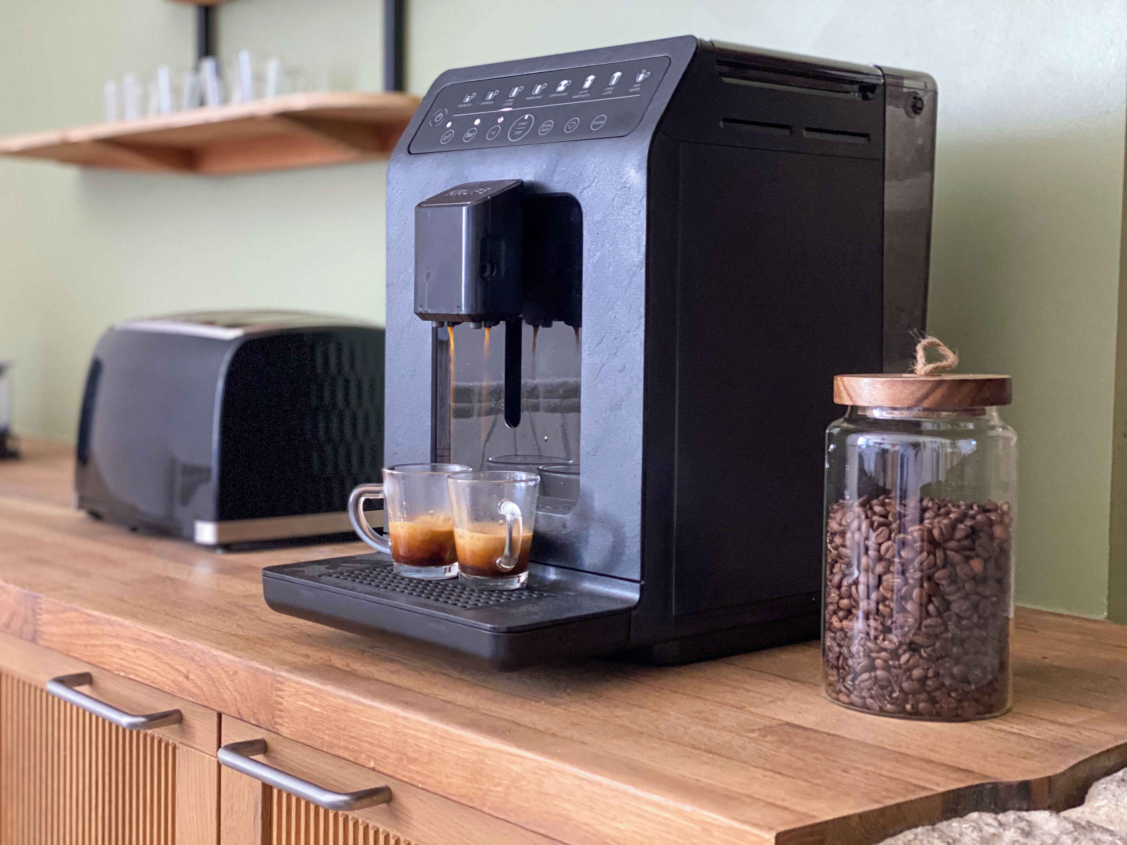 Una cafetera sobre una encimera de madera, con un tarro de vidrio lleno de granos de café a su lado.