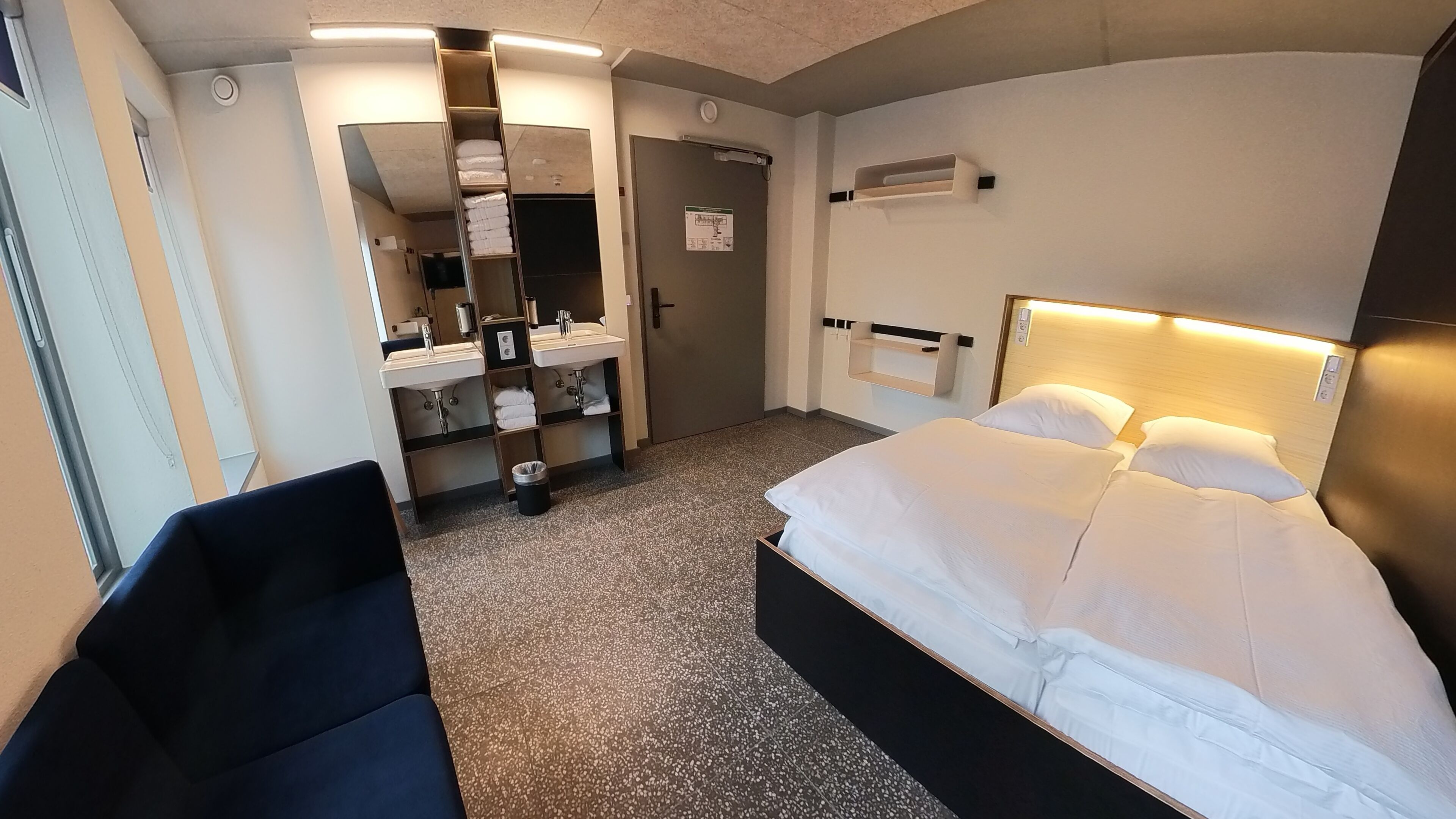 A hotel room with a bed, a desk, a chair, a bathroom with a mirror, and a sink.