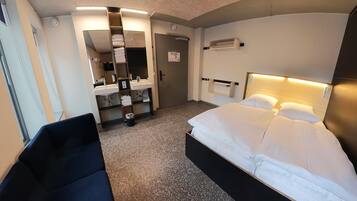 A hotel room with a bed, a desk, a chair, a bathroom with a mirror, and a sink.