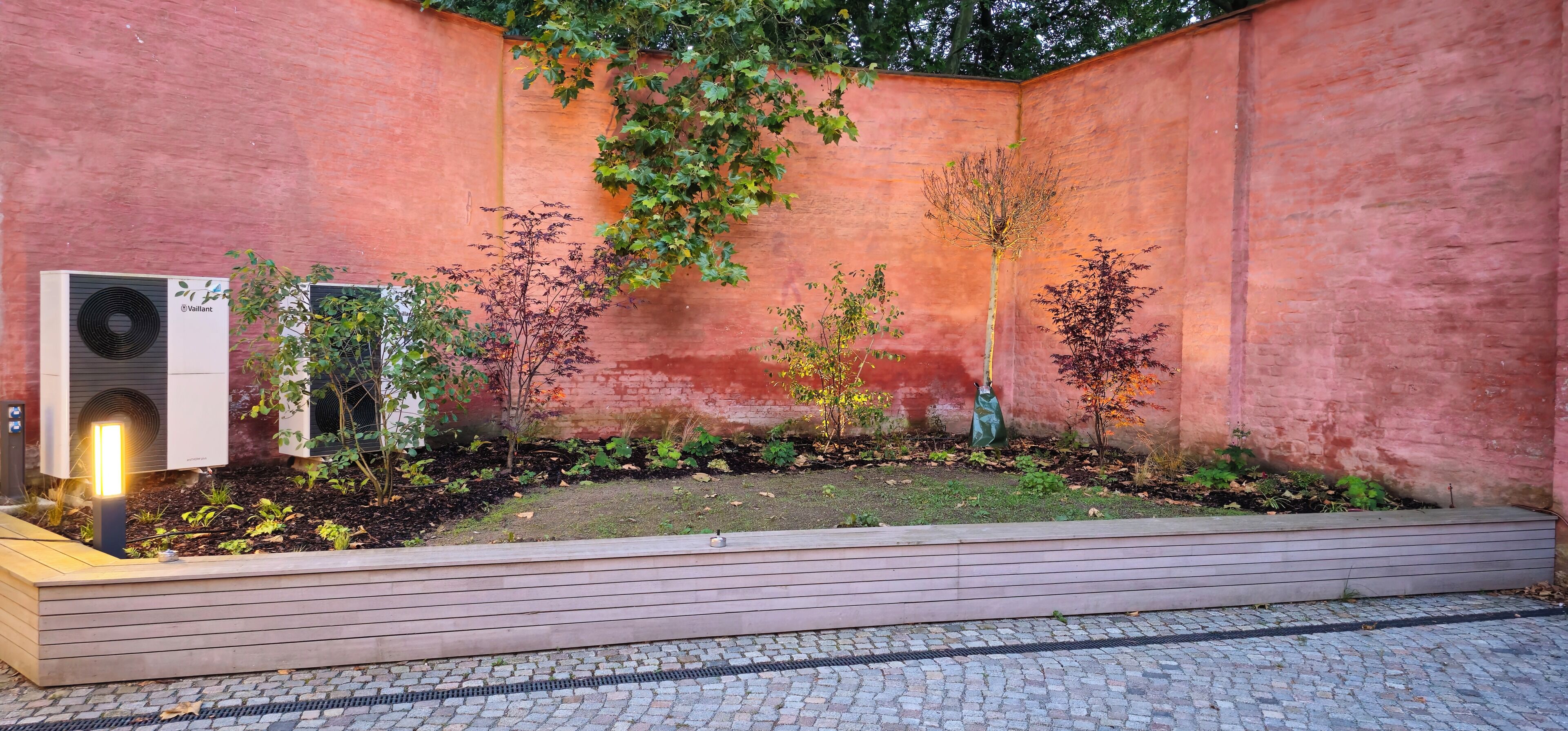 Un patio con pared de ladrillo, un banco de madera y un pequeño jardín con diversas plantas.