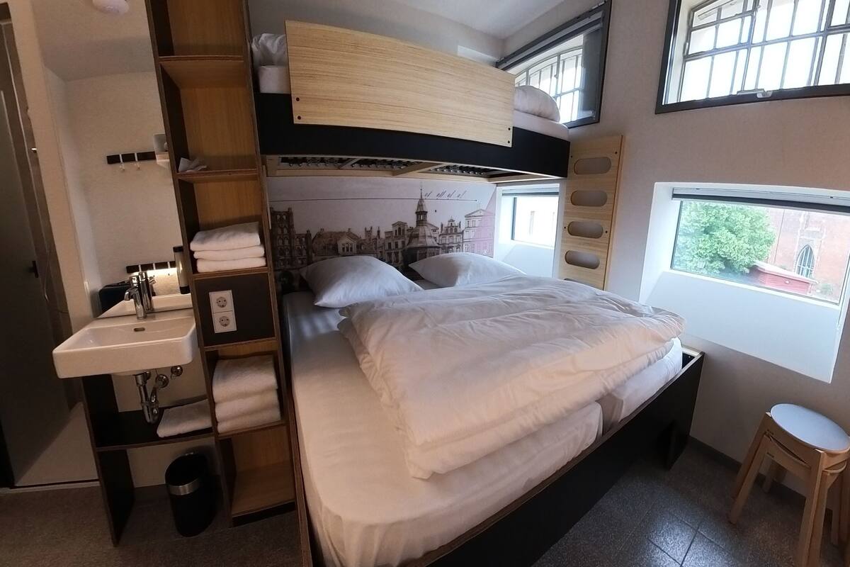 A compact hotel room with a bunk bed, a small sink, a bedside table, and a window with a view of greenery.