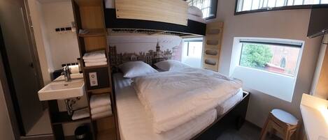 Une chambre d’hôtel compacte avec un lit superposé, un petit lavabo, une table de chevet et une fenêtre donnant sur un espace verdoyant.