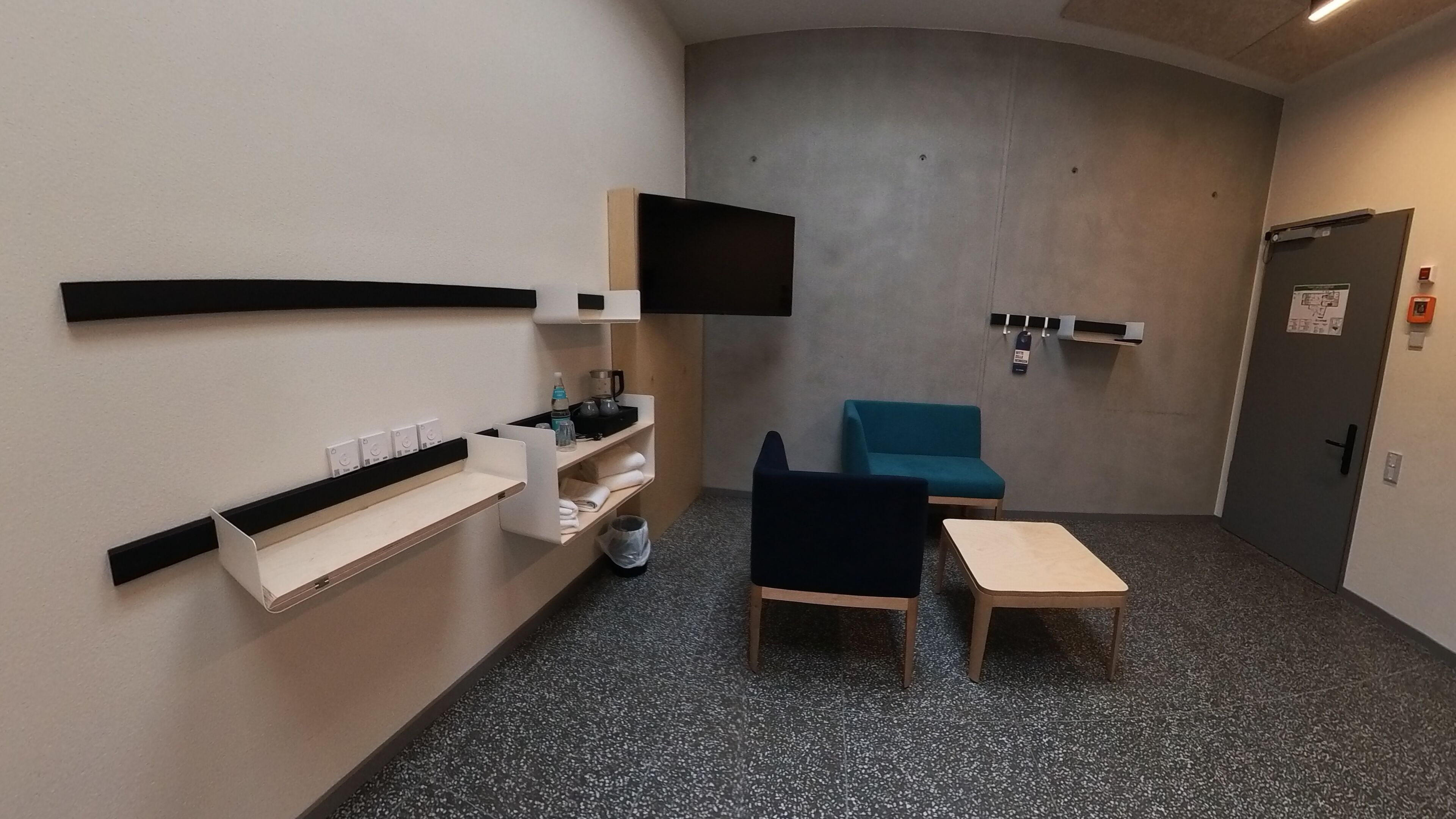 Una habitación pequeña y moderna con una televisión de pantalla plana en la pared, un sofá azul, una mesita y una silla blanca.