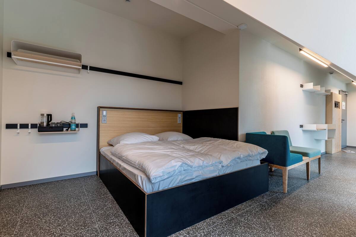 A modern hotel room with a large bed, a desk, a chair, and a small shelf with toiletries.