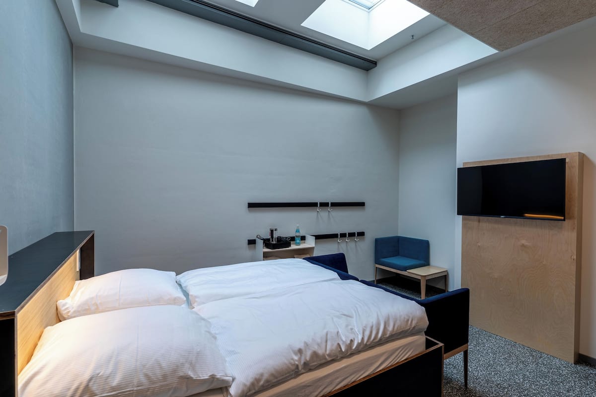 A modern hotel room with a skylight, a bed, a desk, a chair, and a TV mounted on the wall.