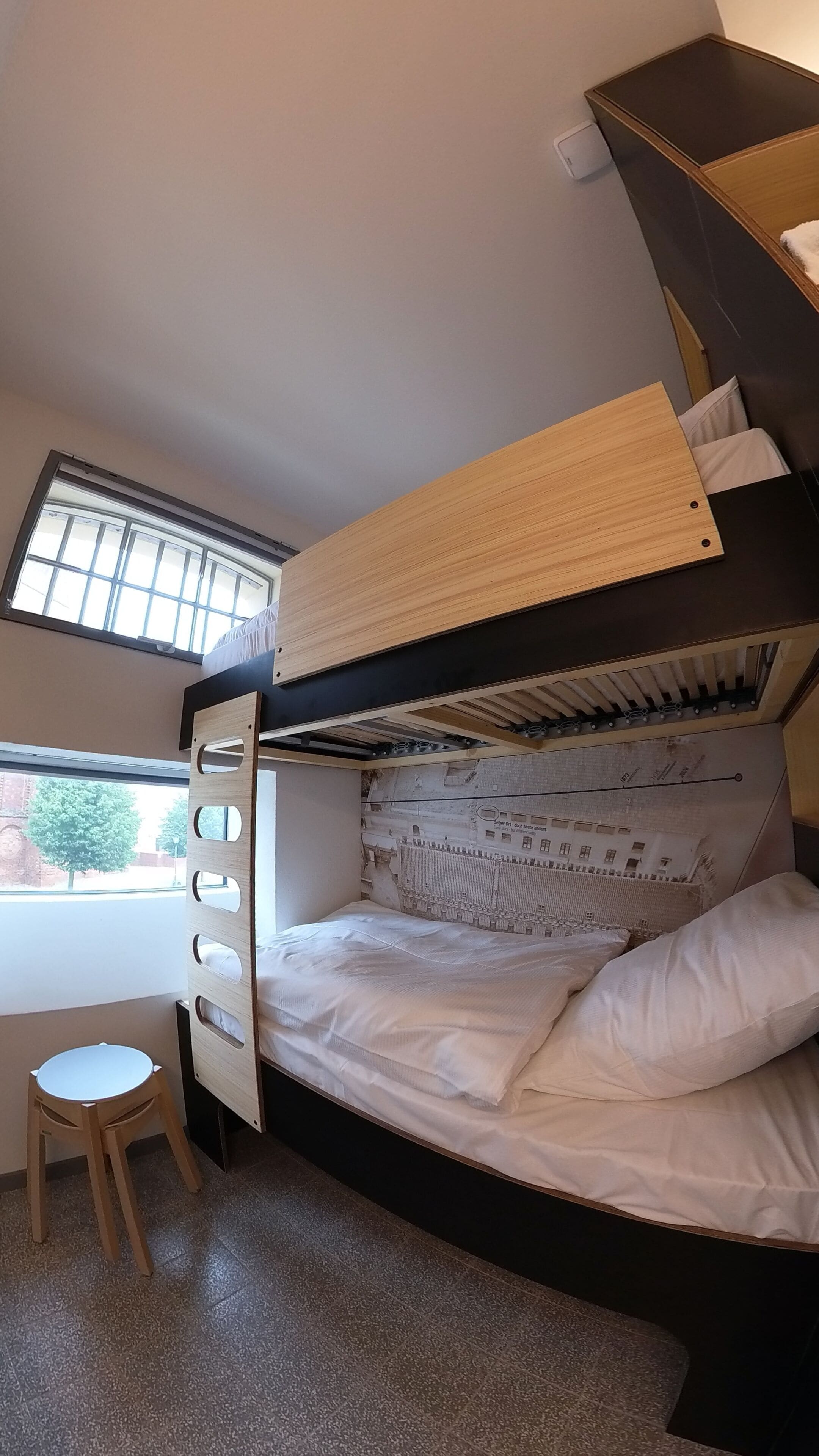 Een stapelbed met een bureau en stoel, een raam met zonwering, en uitzicht op een gebouw.