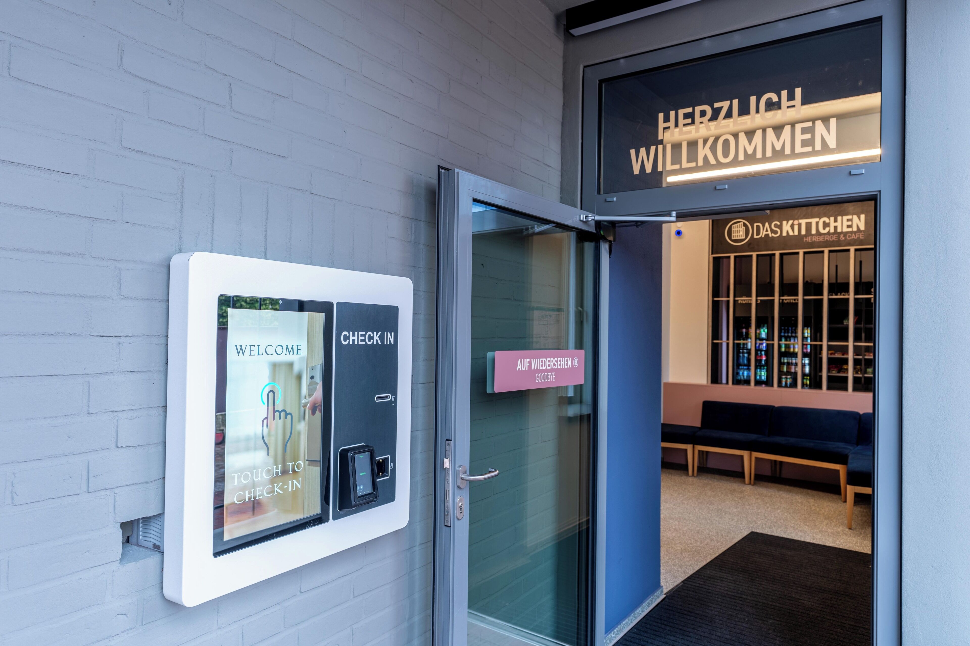 A hotel entrance with a digital check-in kiosk, a welcome sign, and a seating area inside.