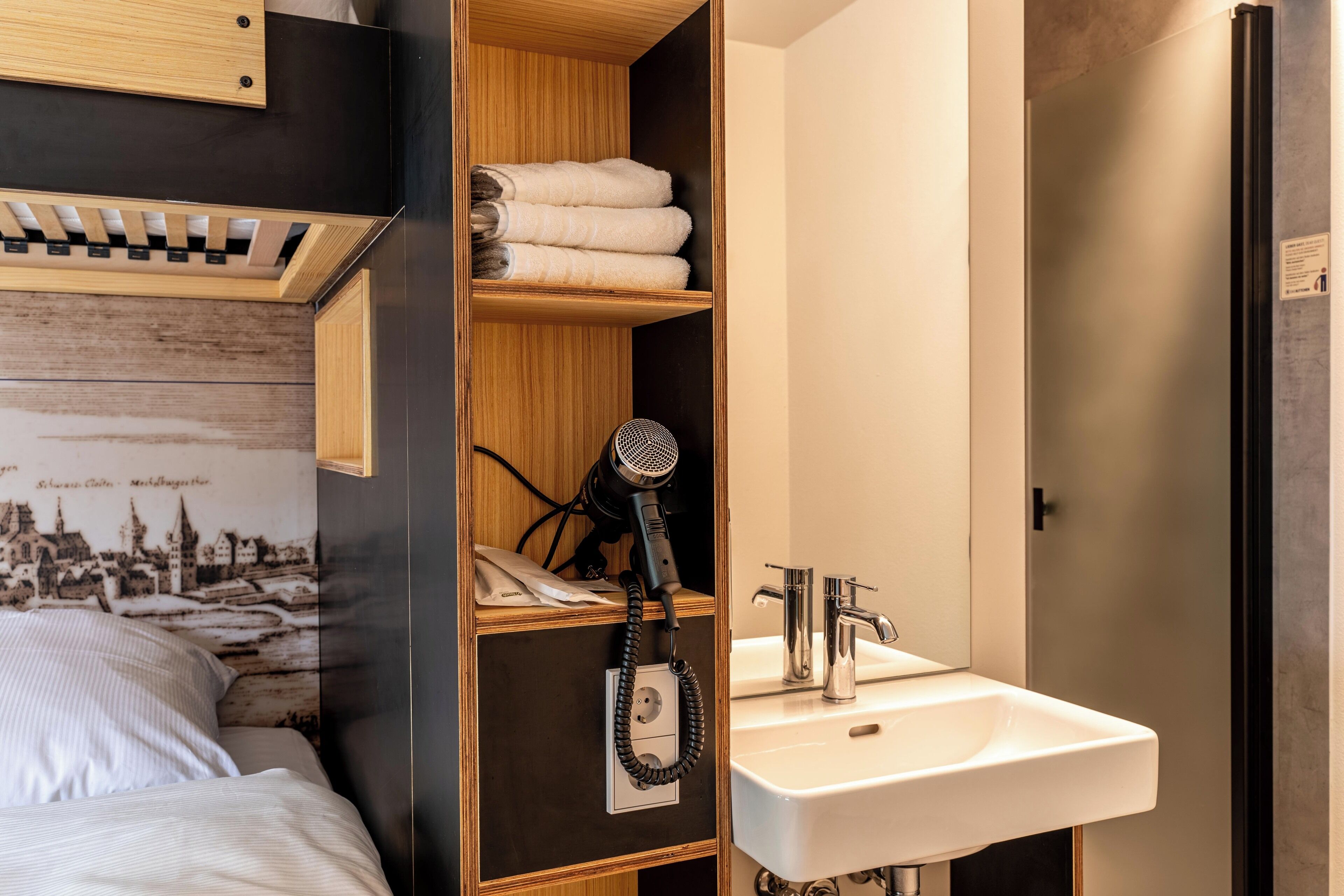 A hotel room with a bed, a wooden shelf with towels, a hairdryer, and a sink.