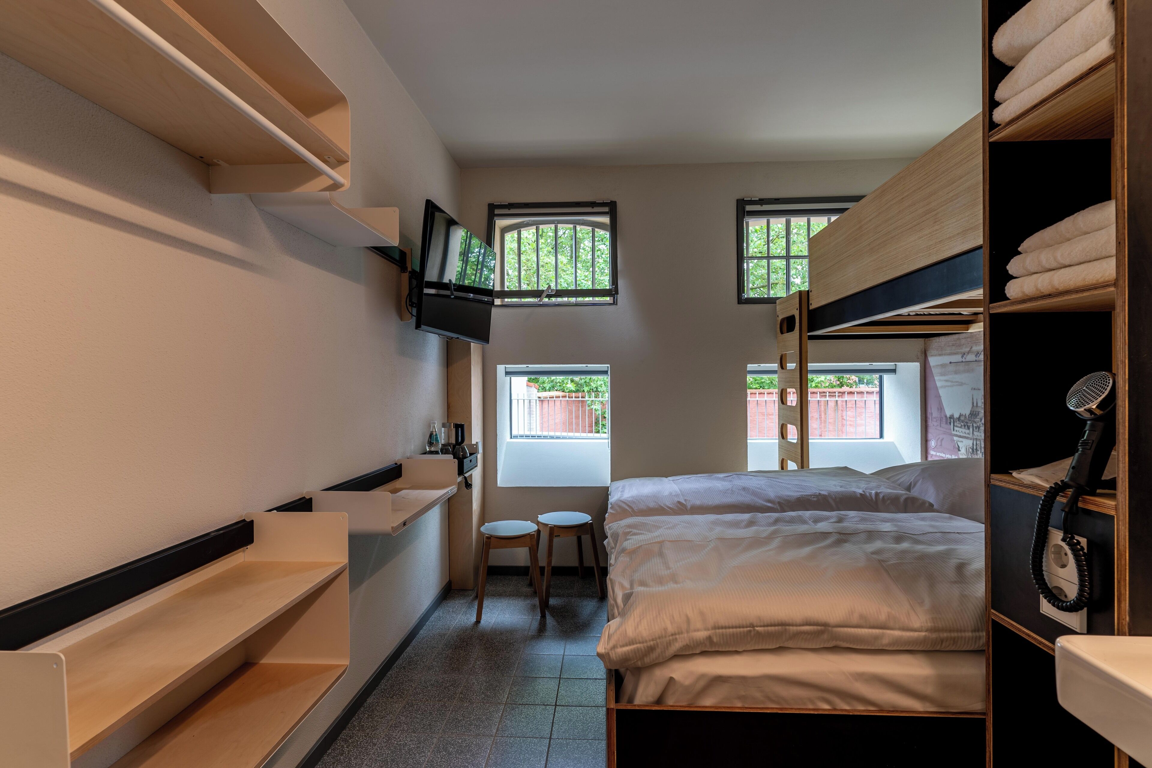 A compact hotel room with a bed, a TV mounted on the wall, a small bathroom with a sink and toilet, and two windows providing natural light.