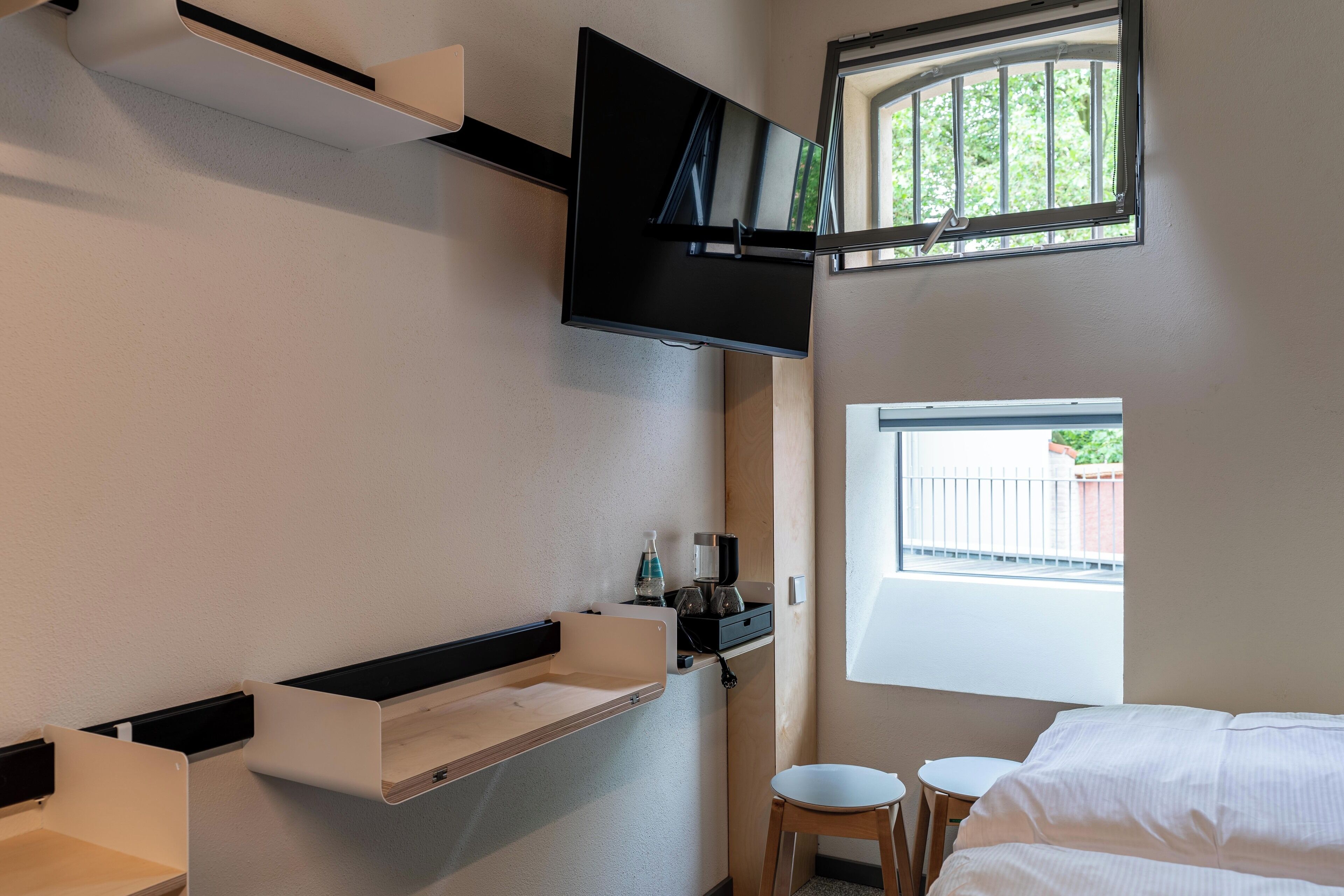 A modern hotel room with a flat-screen TV mounted on the wall, a small window with a view of the outside, and a bed with white bedding.