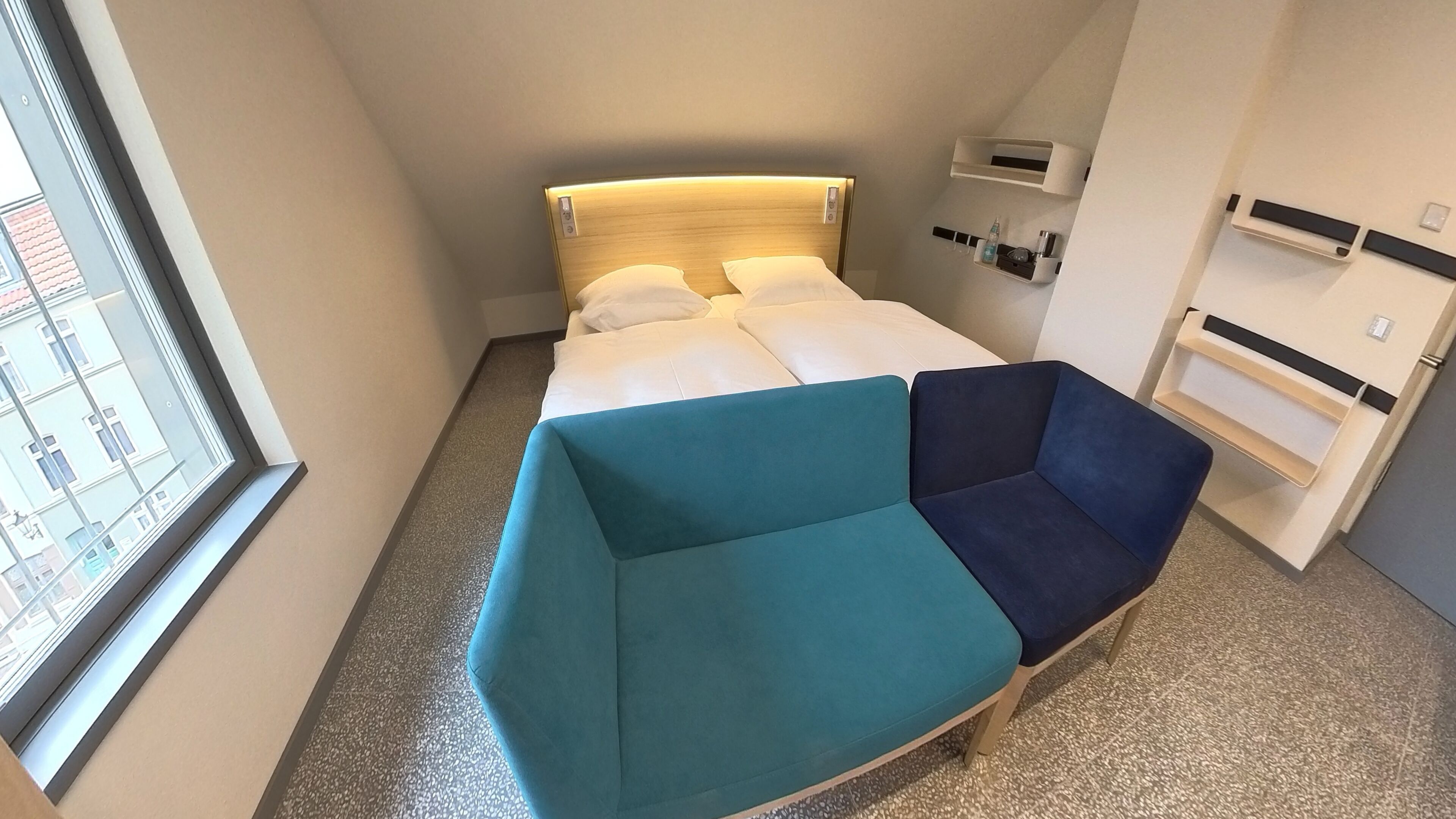Una habitación de hotel con una cama, un sofá azul verdoso y azul marino, una mesita y una ventana.