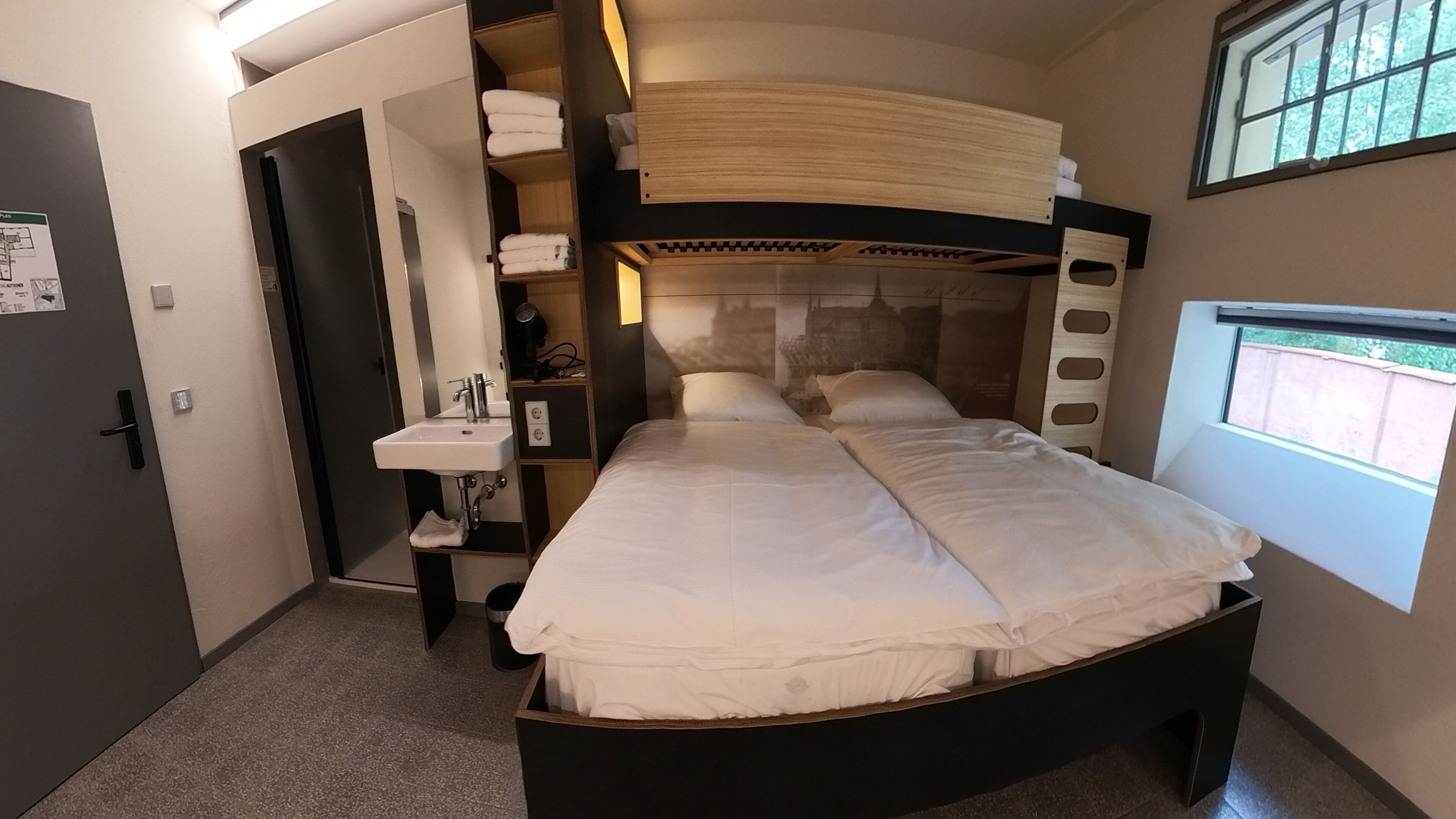 A bedroom with a bunk bed, a bedside table, a sink, and a window with a view.