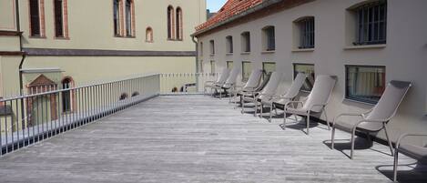 Ein Balkon mit Liegestühlen, einem Geländer und einem Gebäude mit Rundbogenfenstern und einem Ziegeldach.