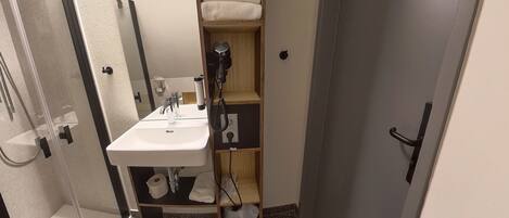 A compact bathroom with a white sink, a towel rack, a shower area, and a shelf with towels.