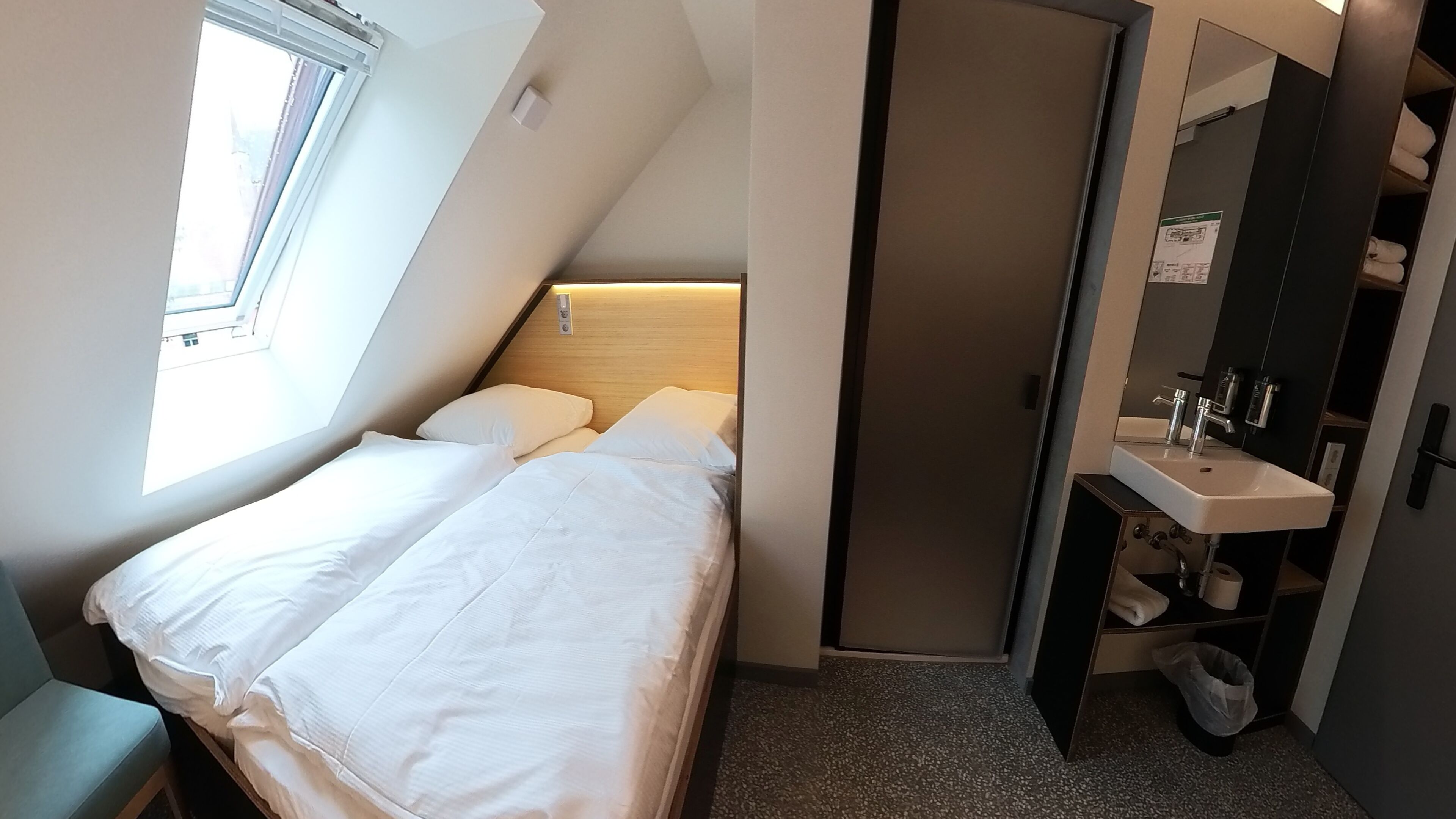 Quarto de hotel pequeno, com cama, janela, banheiro com pia e espelho, e porta que dá acesso a outro cômodo.