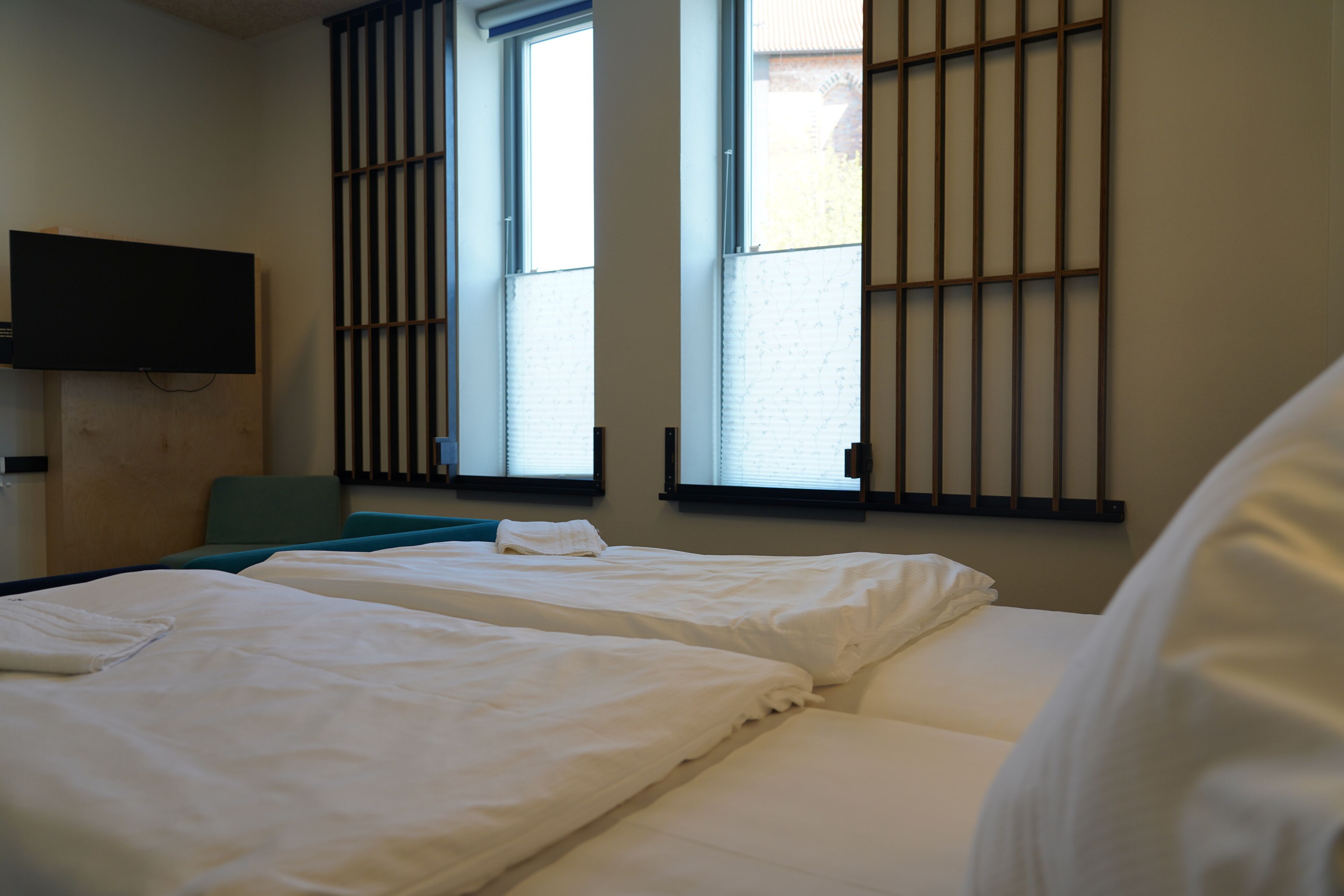 A hotel room with two beds, a television mounted on the wall, and windows with vertical blinds.