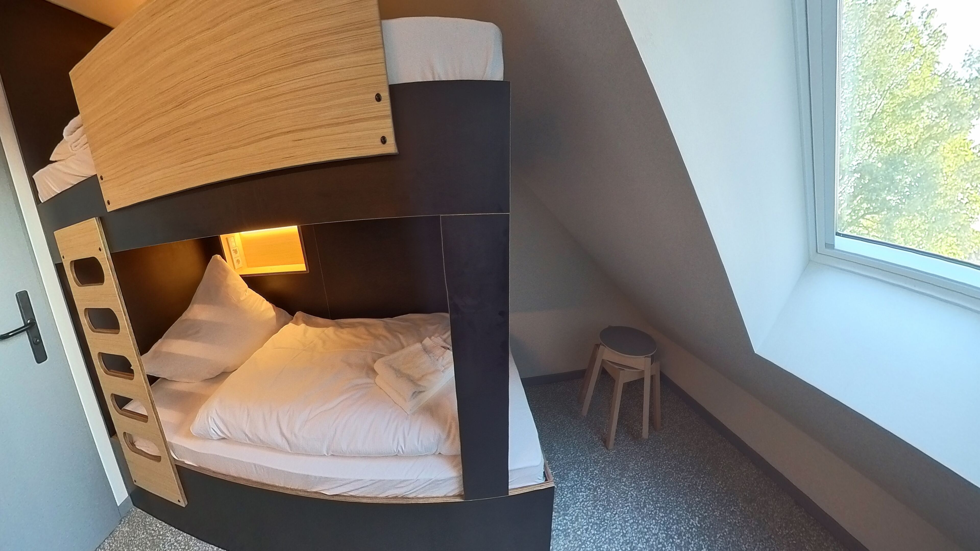 Ein Stockbett mit Holzvorhang, ein kleiner Hocker und ein Fenster mit Blick auf Bäume.