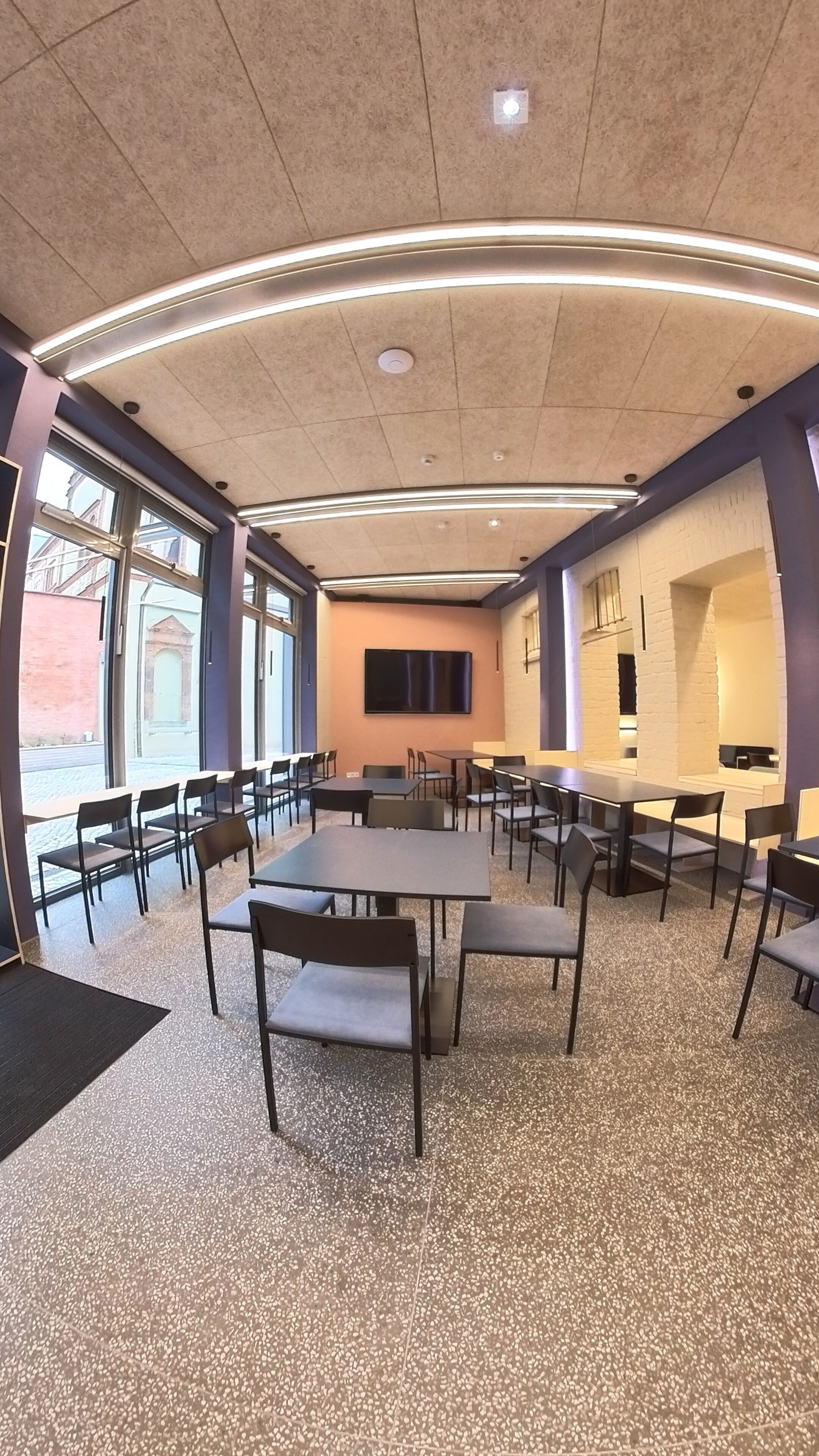 Un salón de conferencias con mesas y sillas, ventanas amplias y un televisor de pantalla plana en la pared.