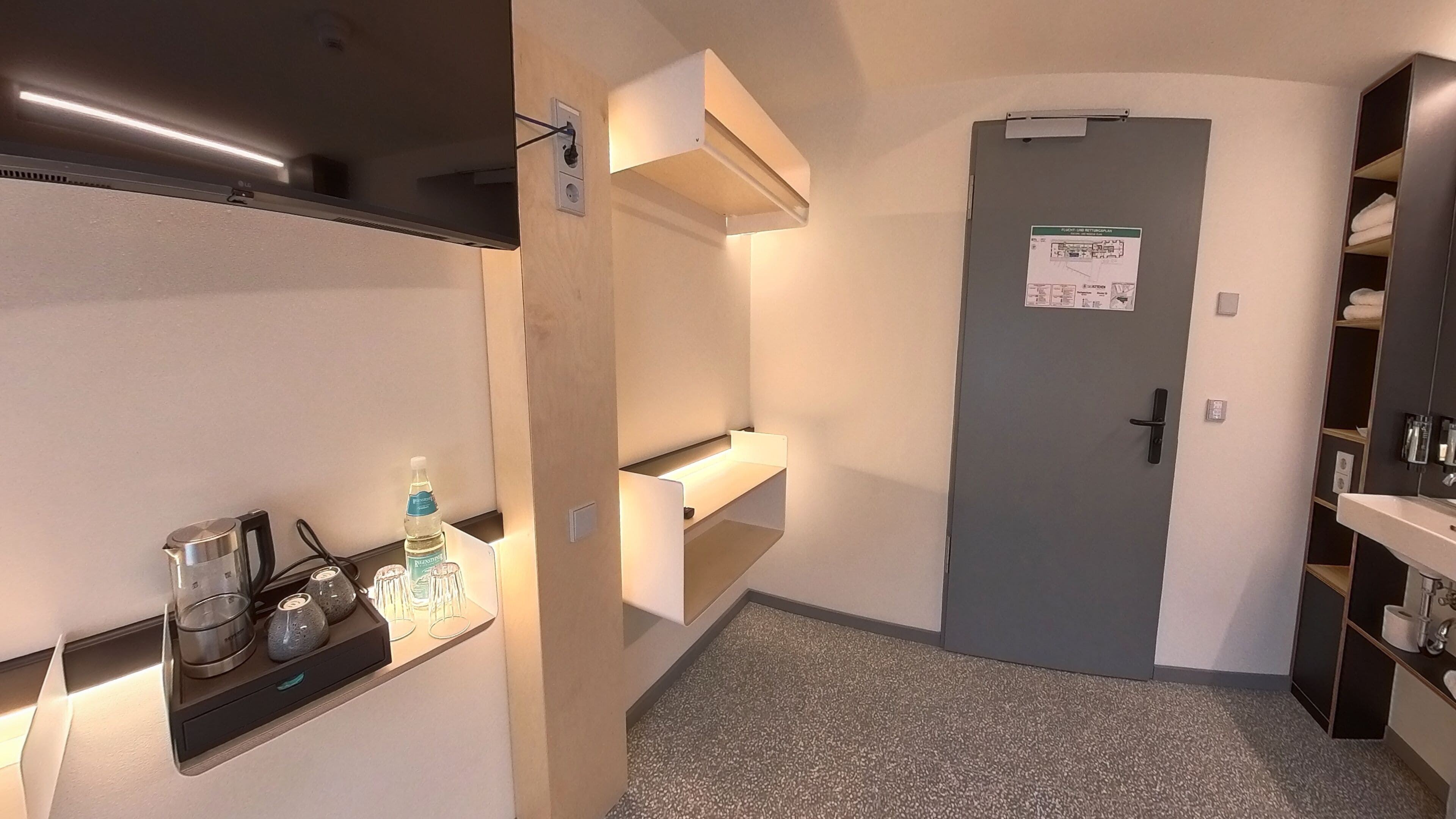 Une chambre d’hôtel équipée d’une kitchenette, d’un petit coin salon avec une table et des chaises, d’une porte avec une pancarte et d’une armoire avec des étagères.