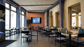 A modern dining area with tables and chairs, a television screen, and large windows.