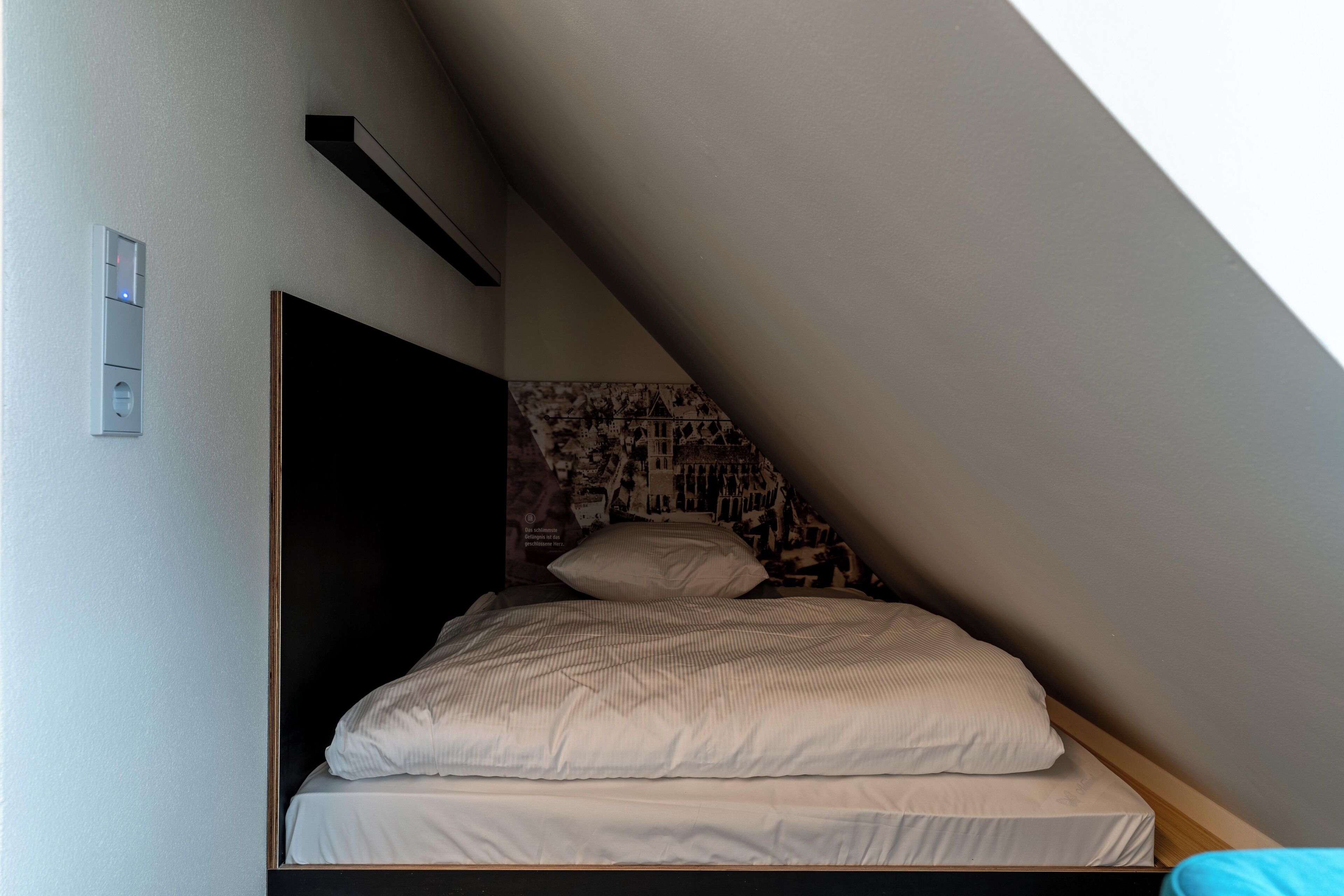 A small, single-bed room with a sloped ceiling, a built-in headboard, and a patterned wall.