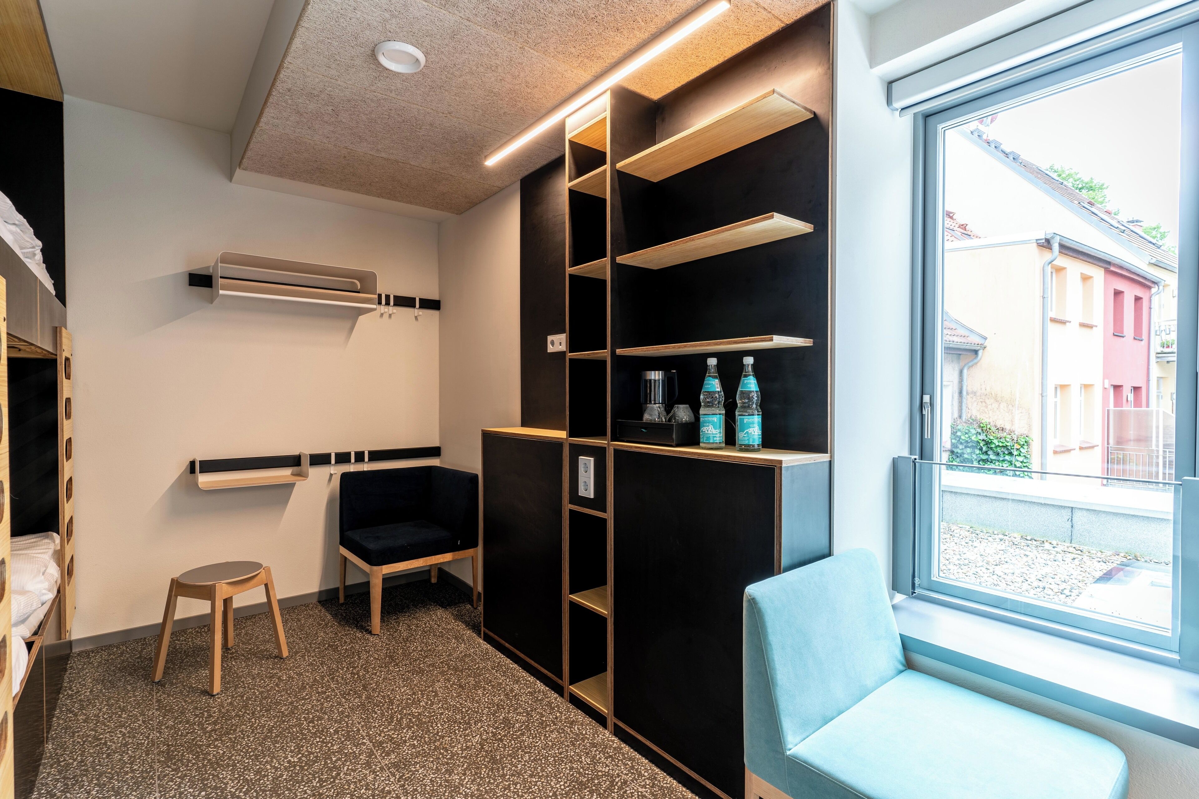 A modern hotel room with a large window, a black and gold storage unit, a blue chair, and a small wooden stool.