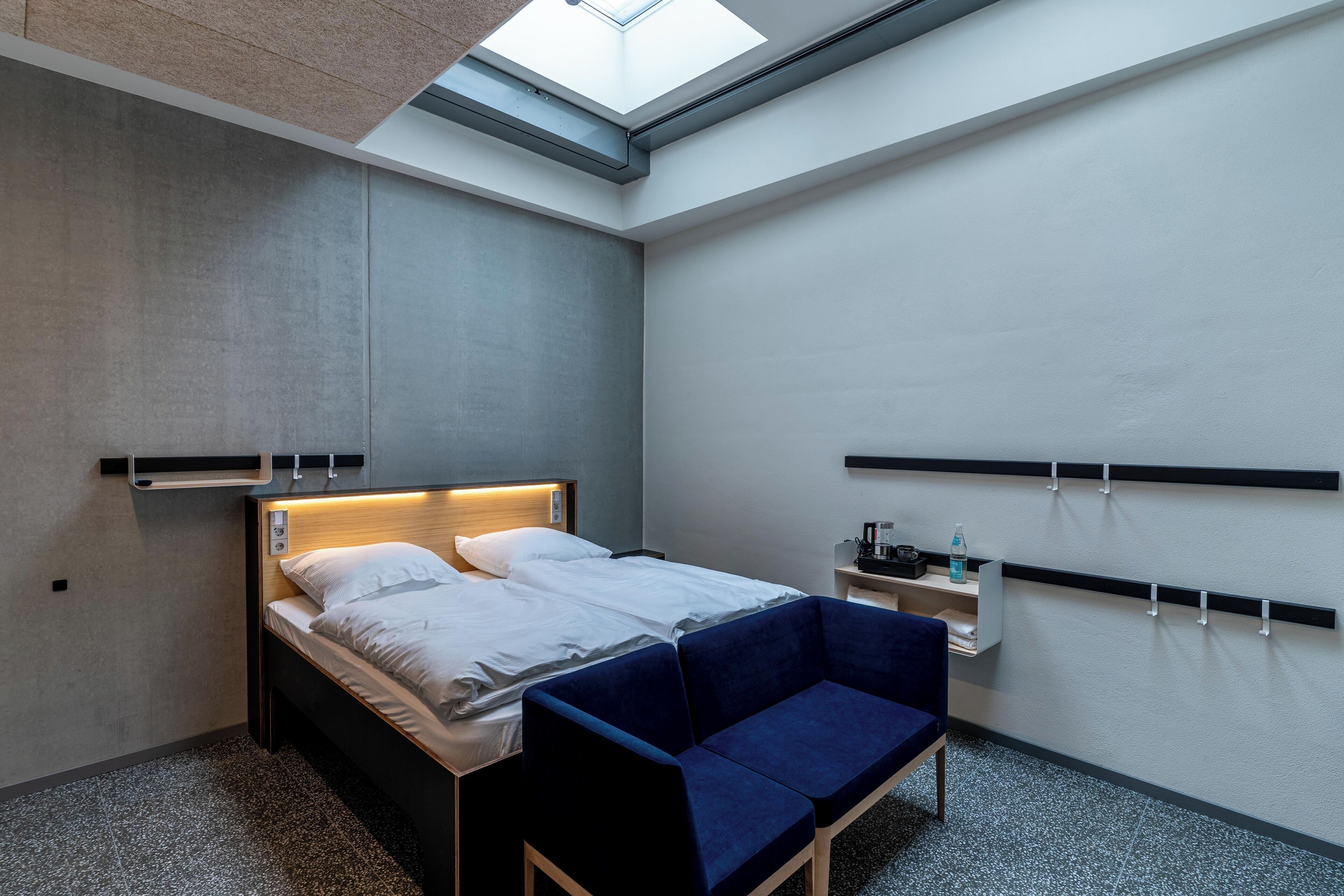 A modern hotel room with a skylight, a bed with white linens, a blue sofa, and a small bedside table with a bottle and a cup.