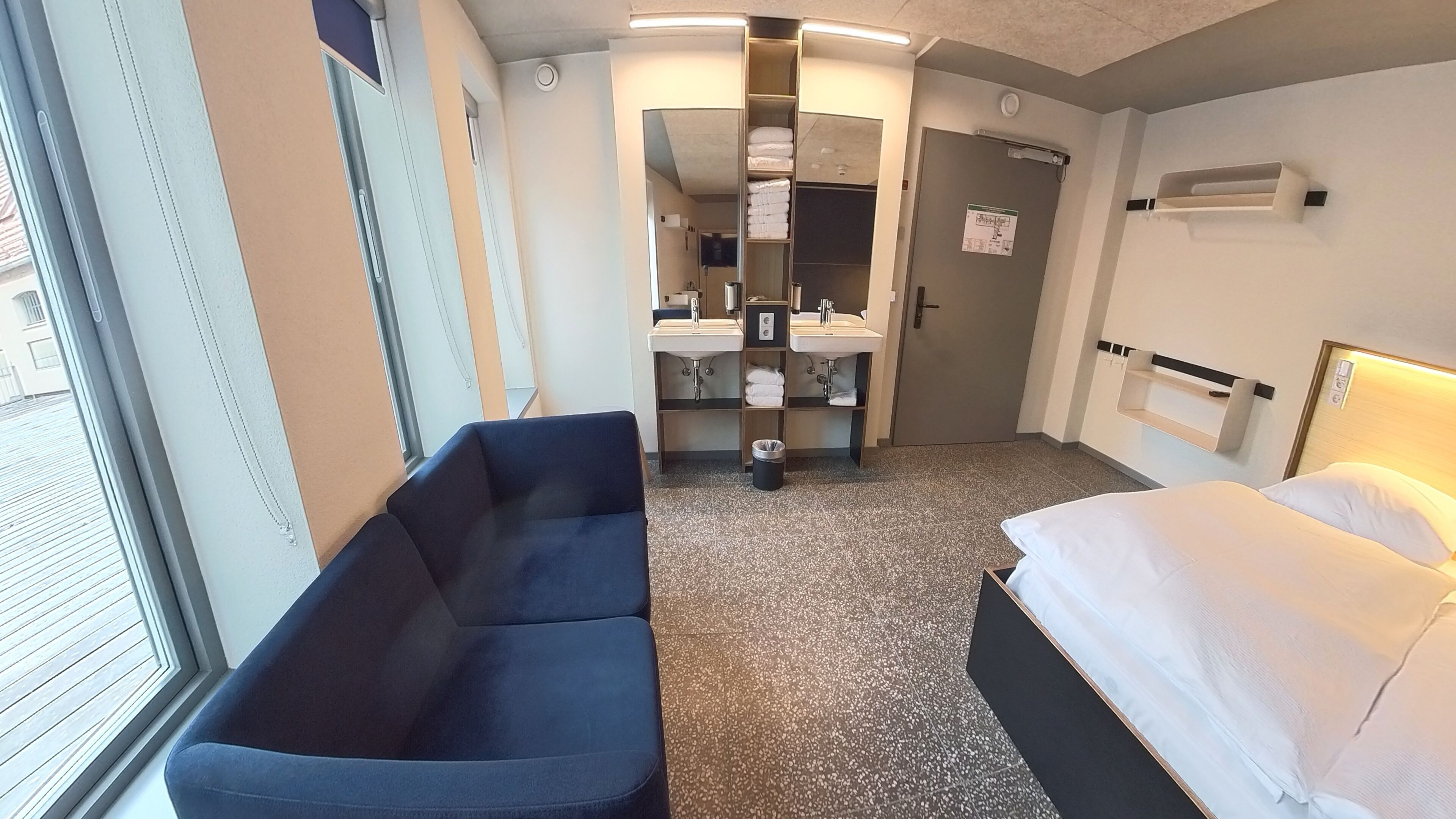 A hotel room with a bed, a sofa, a bathroom with two sinks, and a trash bin.