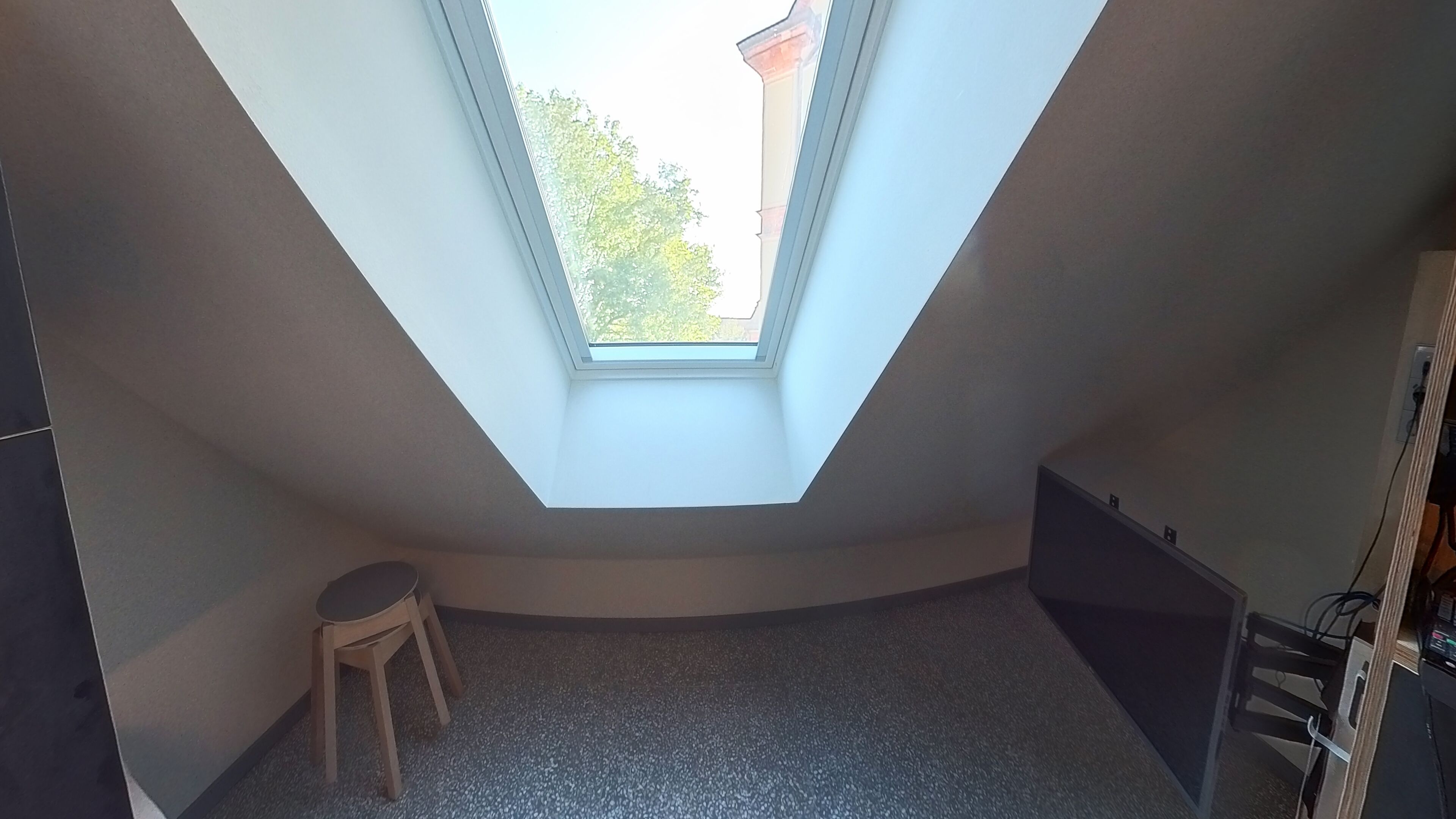 Une pièce avec un plafond en pente et un puits de lumière, un petit tabouret et un radiateur.