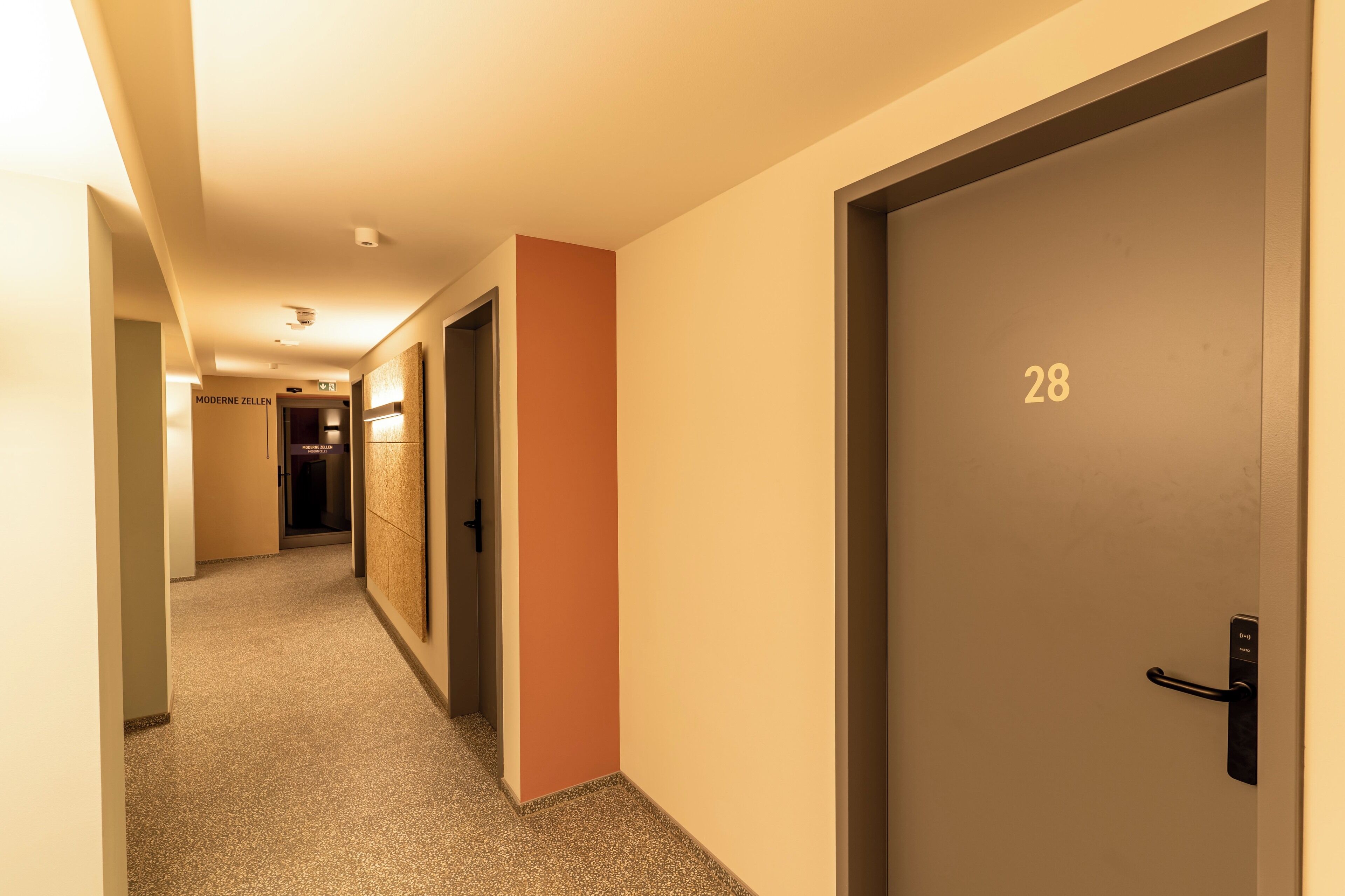 Corredor de hotel com piso carpeteado, uma porta com a inscrição "28" e uma placa com o texto "MODERNE ZELLEN".
