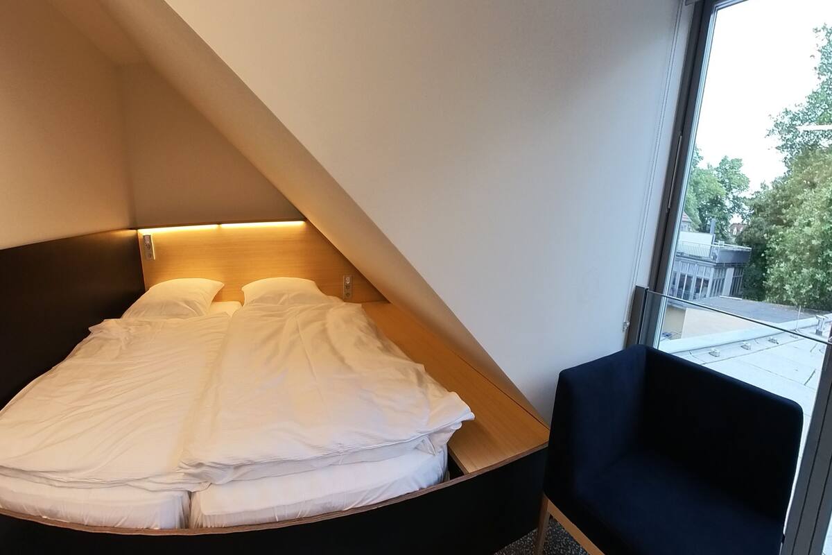 A hotel room with a bed in a corner, a small table, and a chair.