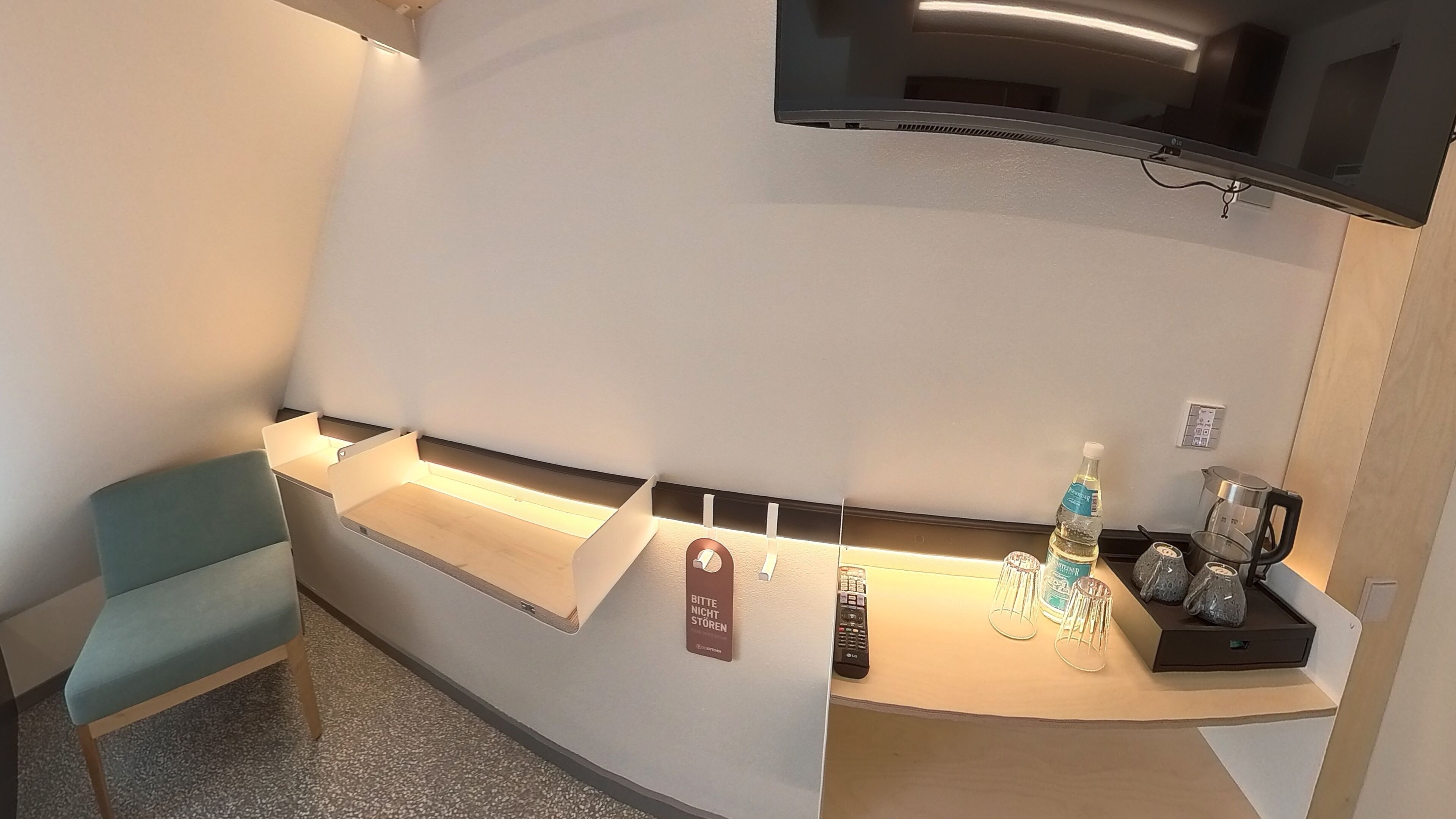 Un espace d’accueil d’hôtel avec un téléviseur à écran plat fixé au mur, un comptoir de réception avec une bouteille d’eau, une bouilloire et une machine à café. Il y a une chaise et un panneau avec du texte accroché au mur.