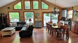 A spacious living area with a high ceiling, a dining table with chairs, a fireplace, and large windows offering views of greenery. - Elberta Bluffs Cottage (Elberta)