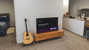 A modern living room with a flat-screen TV on a wooden stand, a guitar, and a kitchen area in the background. - Lakeside apartment, walking distance to foreshore (Kingston)