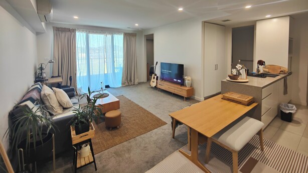 A modern living room with a sofa, coffee table, and TV. There is a dining area with a wooden table and bench. A guitar is leaning against the wall. - Lakeside apartment, walking distance to foreshore (Kingston)