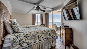Een slaapkamer met een bed, een plafondventilator, een groot raam met gordijnen en uitzicht op de zee.