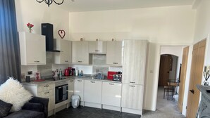 Cozinha moderna com armários brancos, coifa preta e torradeira vermelha. Há uma área de jantar com mesa e cadeiras de madeira, e uma sala de estar com sofá escuro e almofada branca.