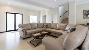 Uma sala de estar espaçosa com um amplo sofá em forma de L bege, uma mesa de centro de madeira e uma escada que leva a outro andar.