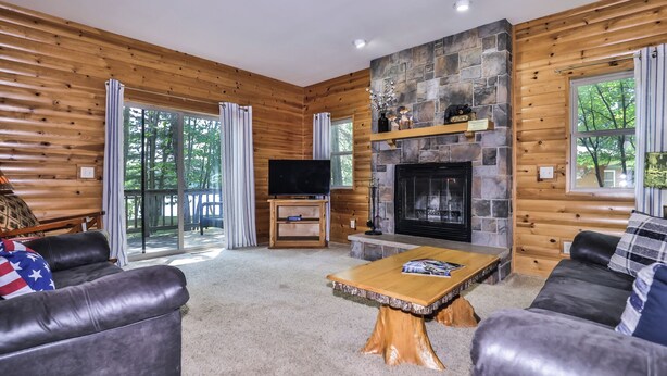 A log cabin interior with a stone fireplace, a television, and a large window overlooking a deck. - Estrold's Resort-Maple Cabin (St.Germain)