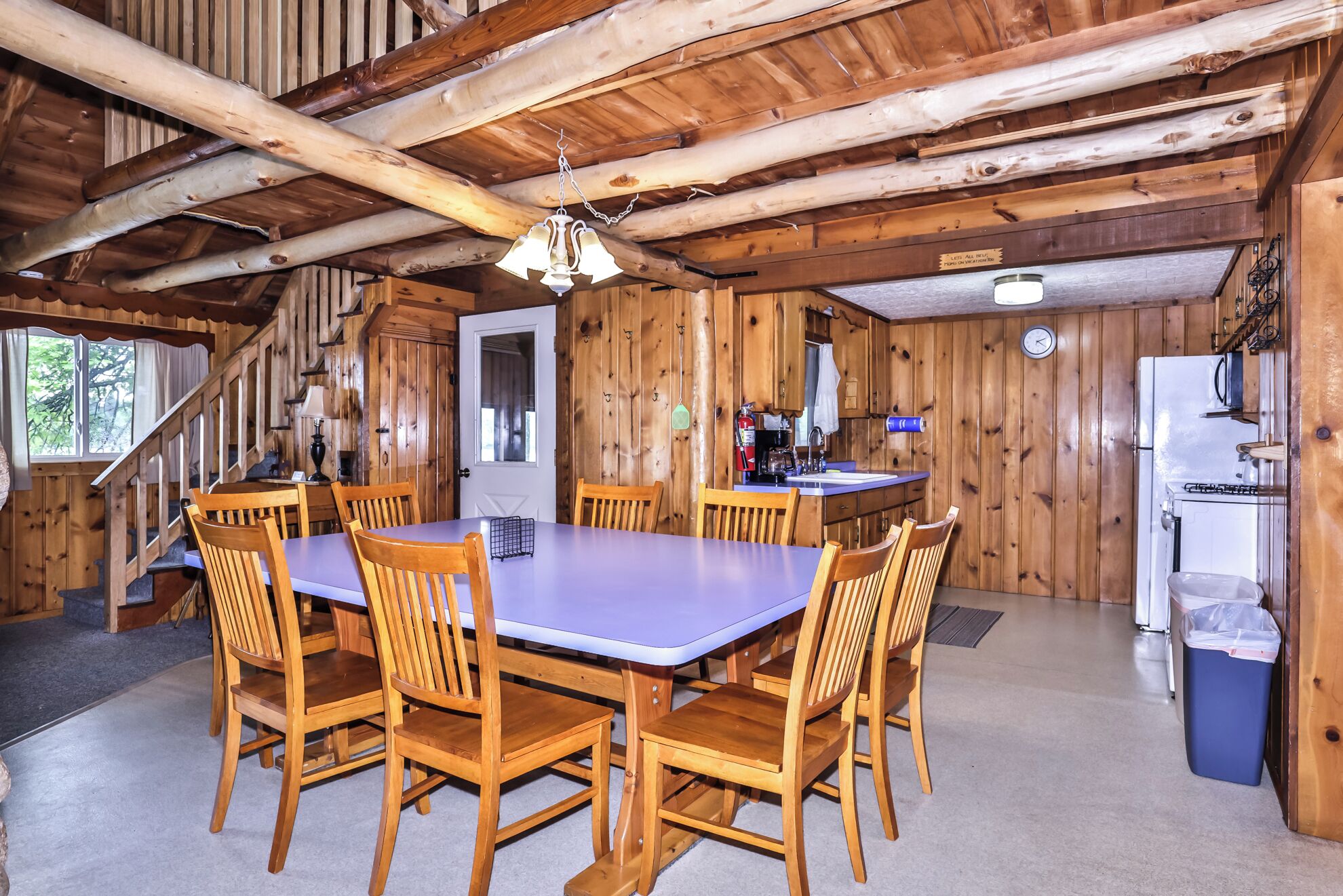 A rustic wooden cabin with a dining area featuring a table and chairs, a kitchen with basic appliances, and a staircase leading to an upper level.