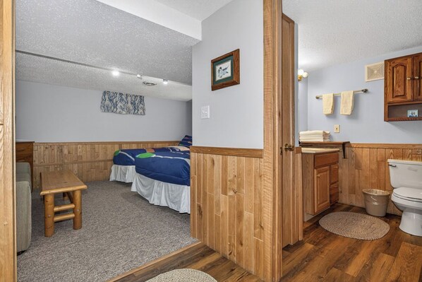 A room with a bed, a small wooden table, a bathroom with a toilet and wooden cabinets, and a wall with a framed picture. - Estrold's Cabin- Red Oak Cabin (St.Germain)