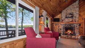 Interior acogedor de una cabaña con chimenea de piedra, sillones tapizados en rojo y un gran ventanal con vista a un lago.