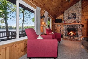 A cozy cabin interior with a stone fireplace, red upholstered chairs, and a large window overlooking a lake.