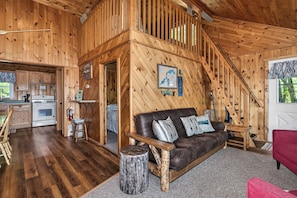 A wooden cabin interior with a kitchen, a dining area, a living room with a sofa, and a staircase.