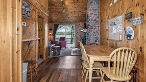 Interior de una cabaña de madera con una mesa de comedor, sillas, una chimenea de piedra y vistas al exterior a través de una puerta.