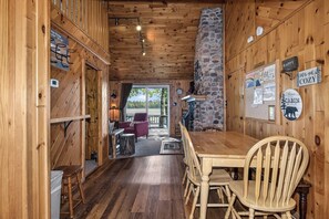 A wooden cabin interior with a dining table, chairs, a stone fireplace, and a view through a door to an outdoor area.