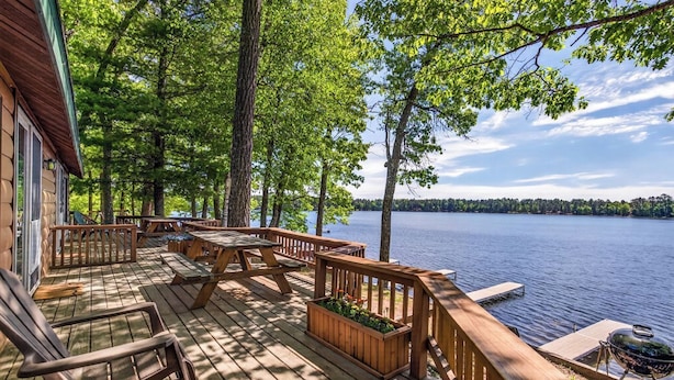 Una terraza de madera con mobiliario exterior, una parrilla y vistas a un lago rodeado de árboles.