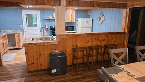 A compact kitchen with wooden cabinets, a microwave, a refrigerator, and a sink. There are stools and a table with a woven placemat.