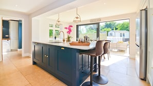 A modern kitchen with a large island, bar stools, pendant lighting, and a view to the outside. - Adgestone Villa (Sandown)